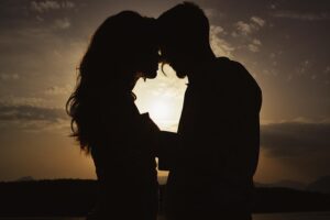 silhouette of a couple standing close, foreheads touching, with the sun setting behind them creating a halo effect