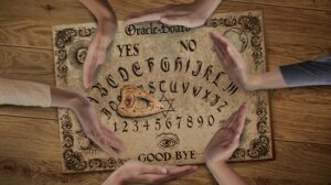 An overhead view of an Oracle board (Ouija board) on a wooden surface, surrounded by several hands with palms open, seemingly interacting with the board