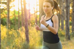 A young woman with a ponytail and headphones is seen jogging through a sunlit forest, holding a phone in her hand
