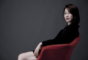A young East Asian woman with dark hair, wearing a black shirt, sits in a red chair and looks over her shoulder at the camera with a subtle smile