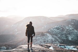 A lone person stands on a snowy mountain peak, gazing out at a vast, snow-dusted mountain range