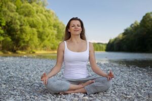 A fair-skinned woman with long brown hair sits cross-legged on smooth river stones by a calm river, eyes closed in peaceful meditation, wearing a white tank top and grey pants.