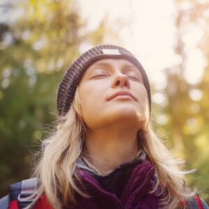 A woman with blonde hair, wearing a beanie and jacket, closes her eyes and enjoys the sunlight while standing in a forest.
