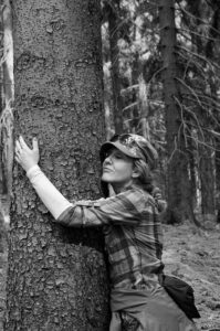 A black and white image of a woman in a plaid shirt and hat, hugging a tree in a forest