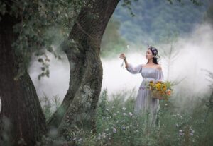 A woman wearing a long, light-colored dress and a floral wreath on her head stands in a misty, green landscape next to a tree