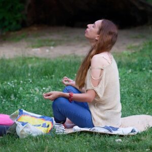 A woman with long brown hair sits cross-legged on a mat in a grassy area, eyes closed and head tilted back