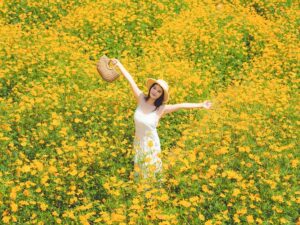 A joyful woman in a white dress and straw hat stands with arms outstretched in the middle of a vast field of bright yellow flowers.