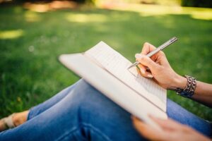 a person's hands writing in a notebook with a pen, while sitting on green grass outdoors