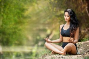 A woman with dark curly hair and olive skin, dressed in a black sports bra and shorts, meditates in a cross-legged position with her eyes closed.